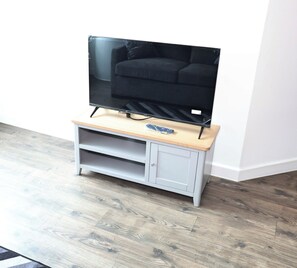A flat-screen TV on a wooden TV stand with a remote control, set against a white wall with a wooden floor. - Cozy Croydon Apartment Retreat (Croydon)