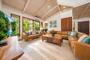 A spacious living area with a high ceiling, a large window with a view of greenery, a sofa set, a coffee table, and a wooden cabinet. - Balinese Poipu Beach Estate -4 ensuites -Full AC -Sleeps 8-16 - Poipu Beach Club (Koloa)