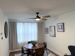 Un comedor con una mesa redonda, sillas, un ventilador de techo y una ventana con cortinas.