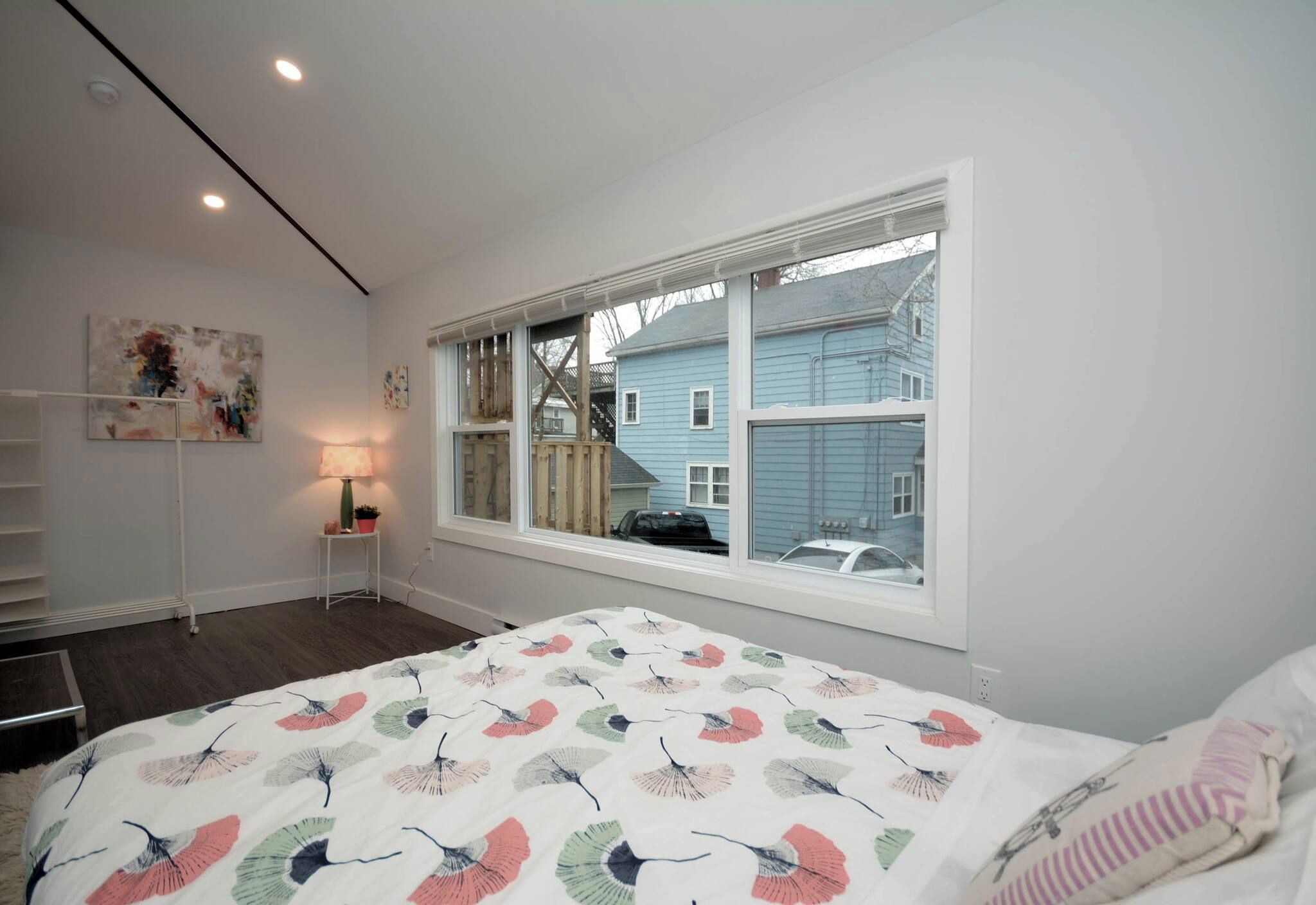 Un dormitorio con un ventanal grande, una cama con cubrecama floral, una mesita de noche con lámpara y un estante en la pared.