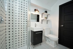 Un baño con un lavamanos blanco, un mueble negro y un inodoro. Hay un espejo sobre el lavamanos y una puerta negra a la derecha.