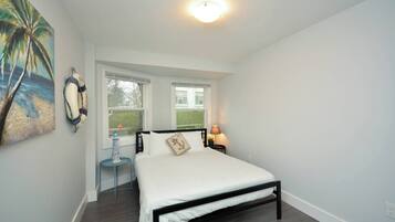 A bedroom with a bed, a nightstand, a lamp, a window, and a painting.