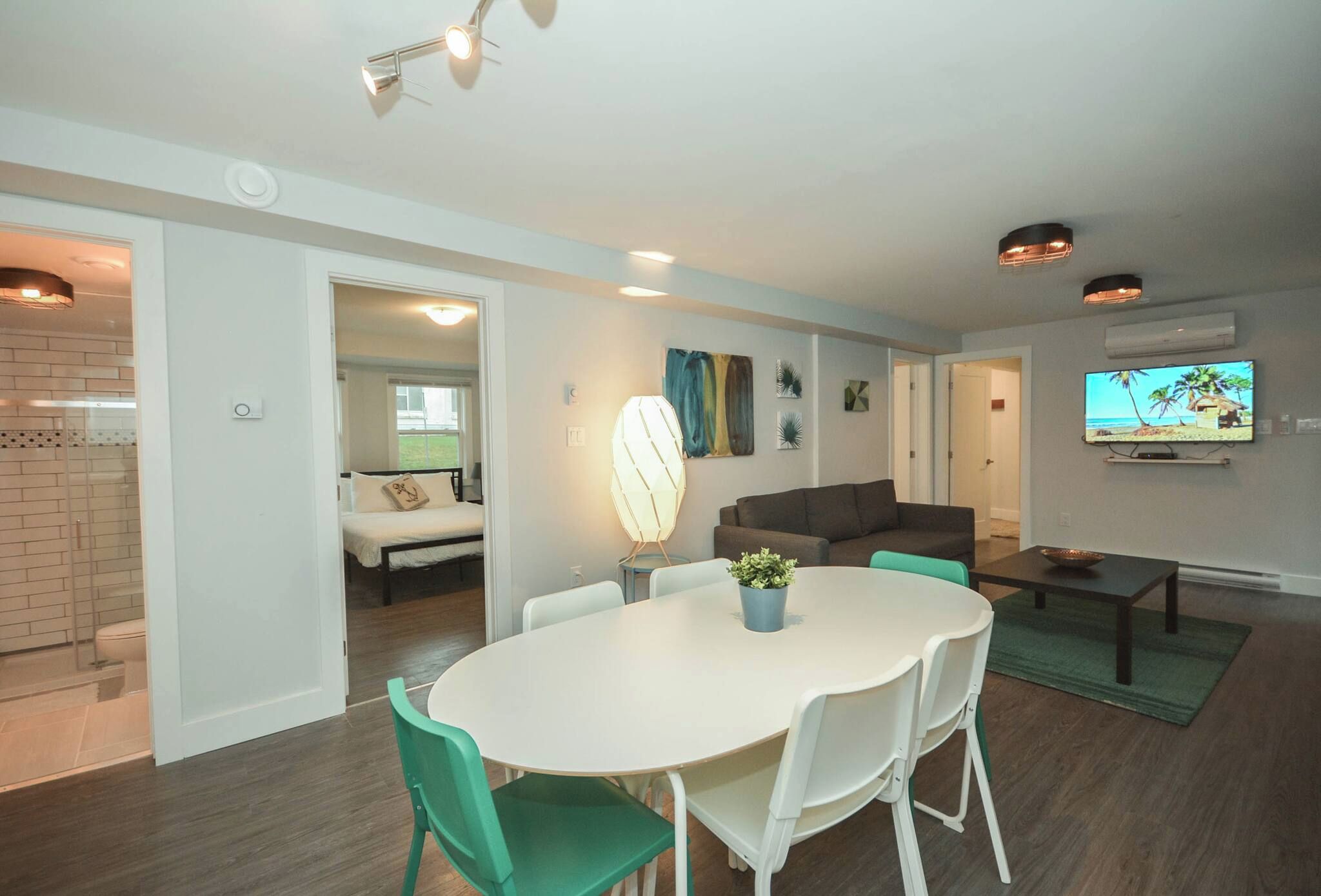 A modern living room with a dining area, a television mounted on the wall, and a visible bathroom with a shower.