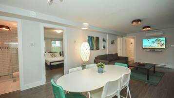 A modern living room with a dining area, a television mounted on the wall, and a visible bathroom with a shower.