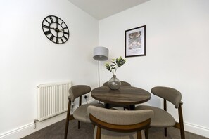 A dining area with a round table, four chairs, a wall clock, a floor lamp, and a framed picture. - The Cosy Pavilion Stylish 2BR Home With Free Street Parking (Durham)