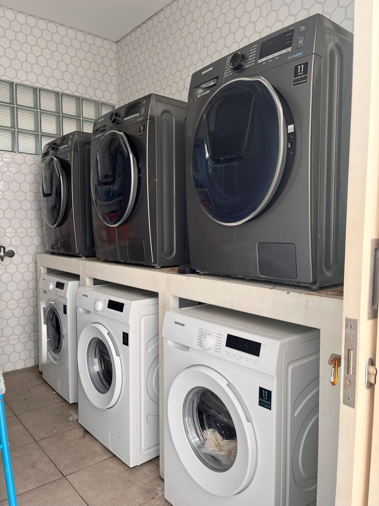 A laundry room with multiple Samsung washing machines, a tiled wall, and a metal rack.