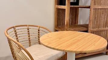 A wicker chair with a cushion, a round wooden table, and a wooden shelving unit with a small electronic device on it.