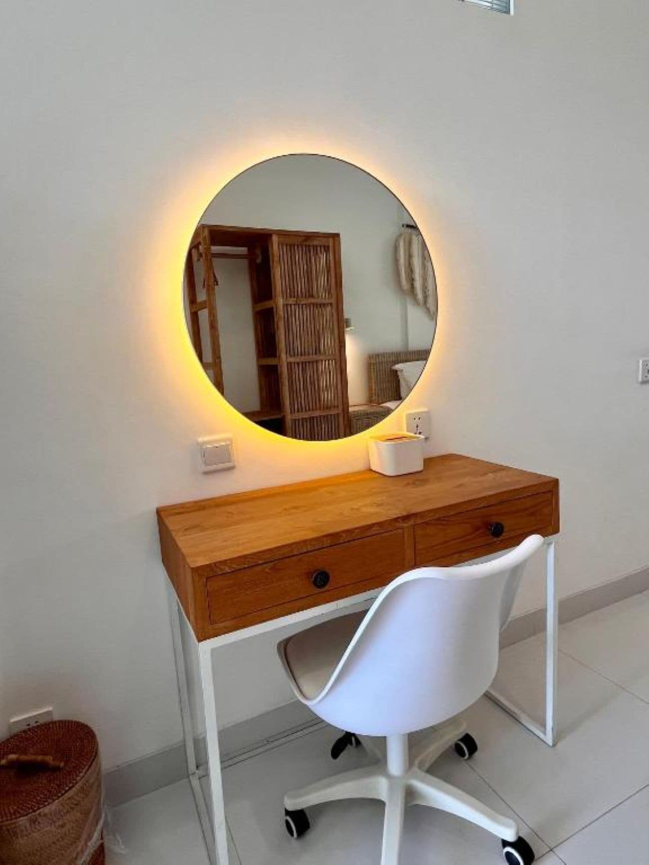 A wooden desk with a round mirror, a white chair, and a small white table with a tissue box.