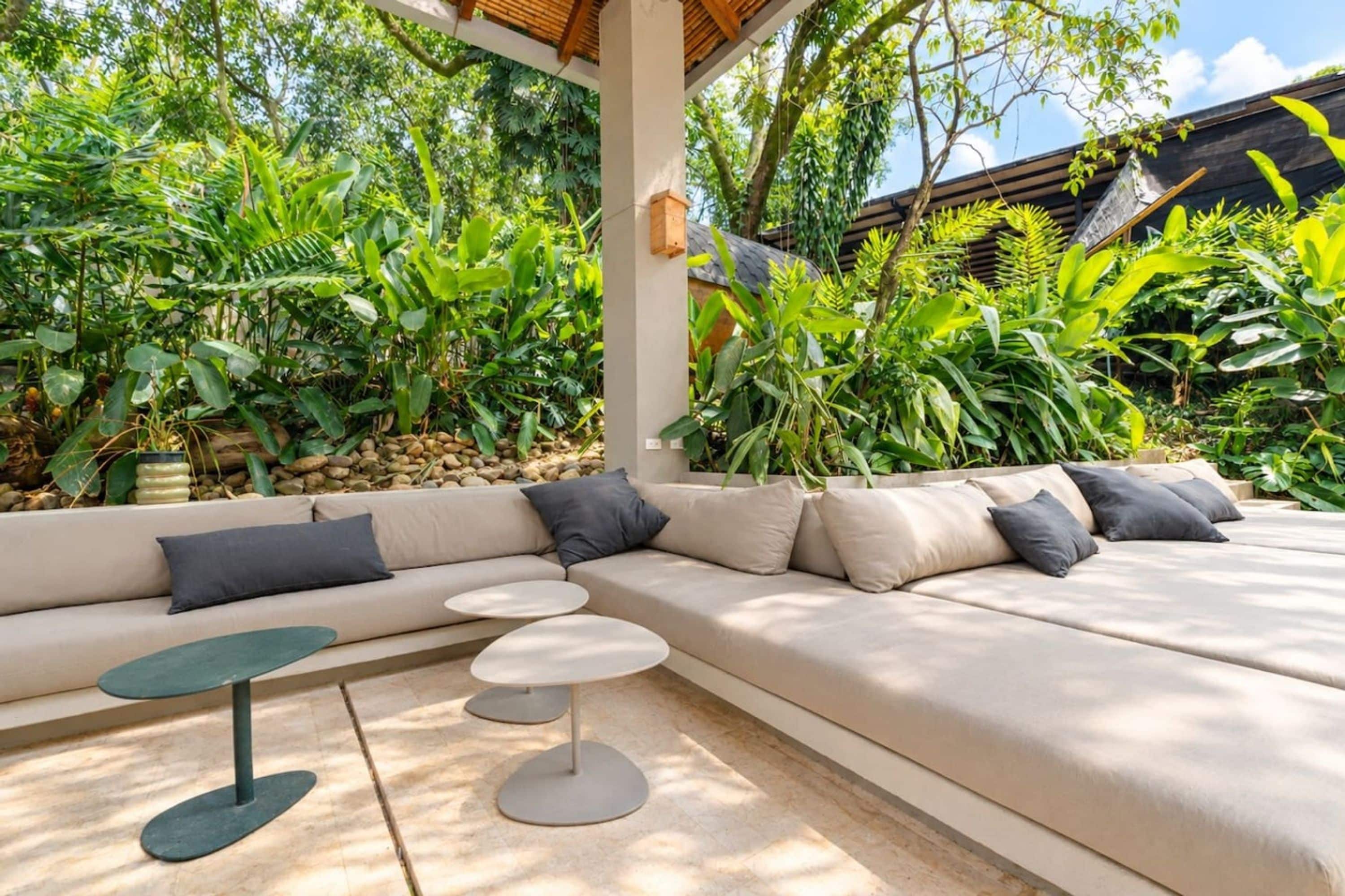 A modern outdoor seating area with a sofa and round tables, surrounded by lush tropical plants.