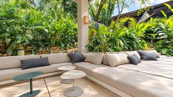 A modern outdoor seating area with a sofa and round tables, surrounded by lush tropical plants.