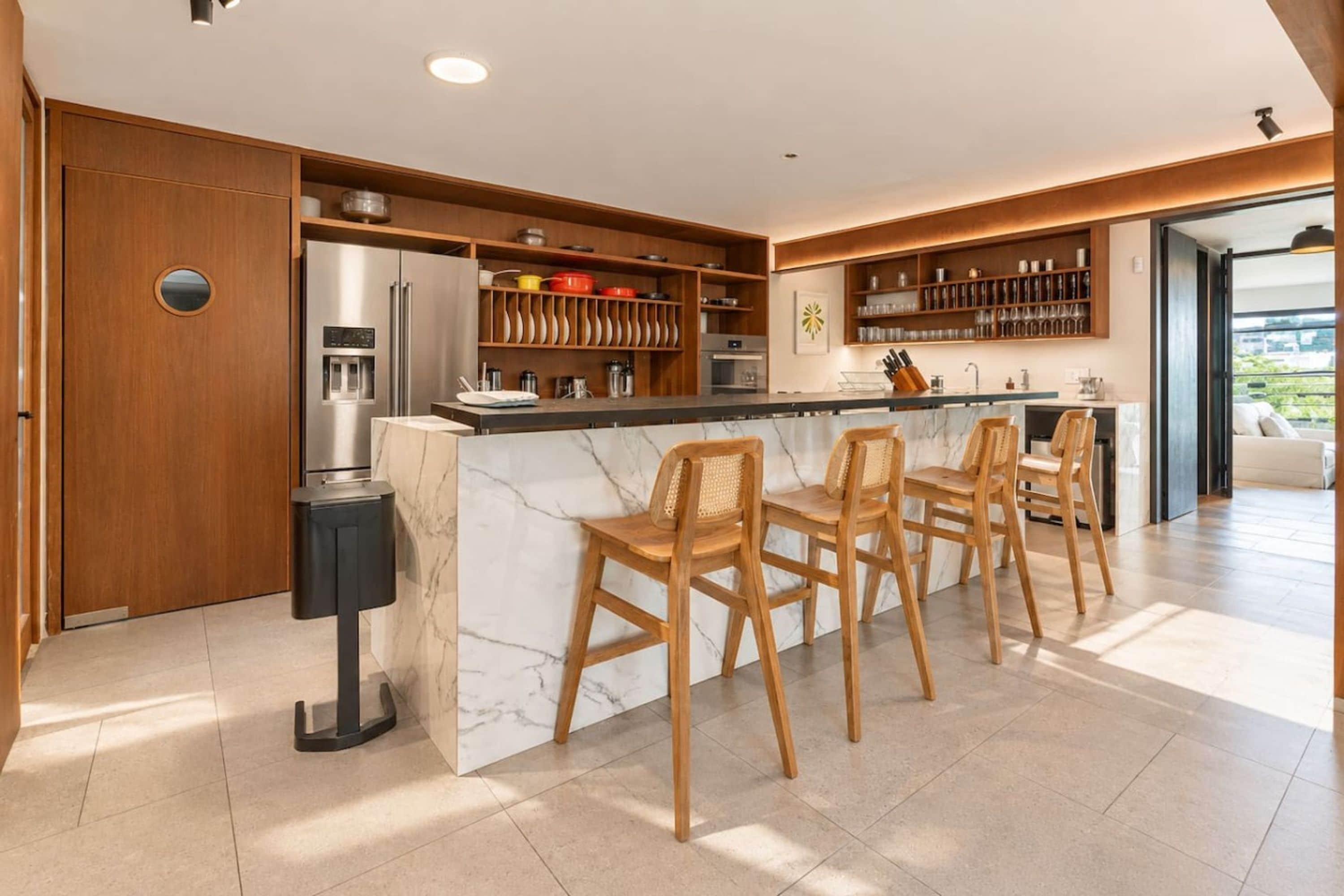 A modern kitchen with a marble island, wooden chairs, and built-in shelving.