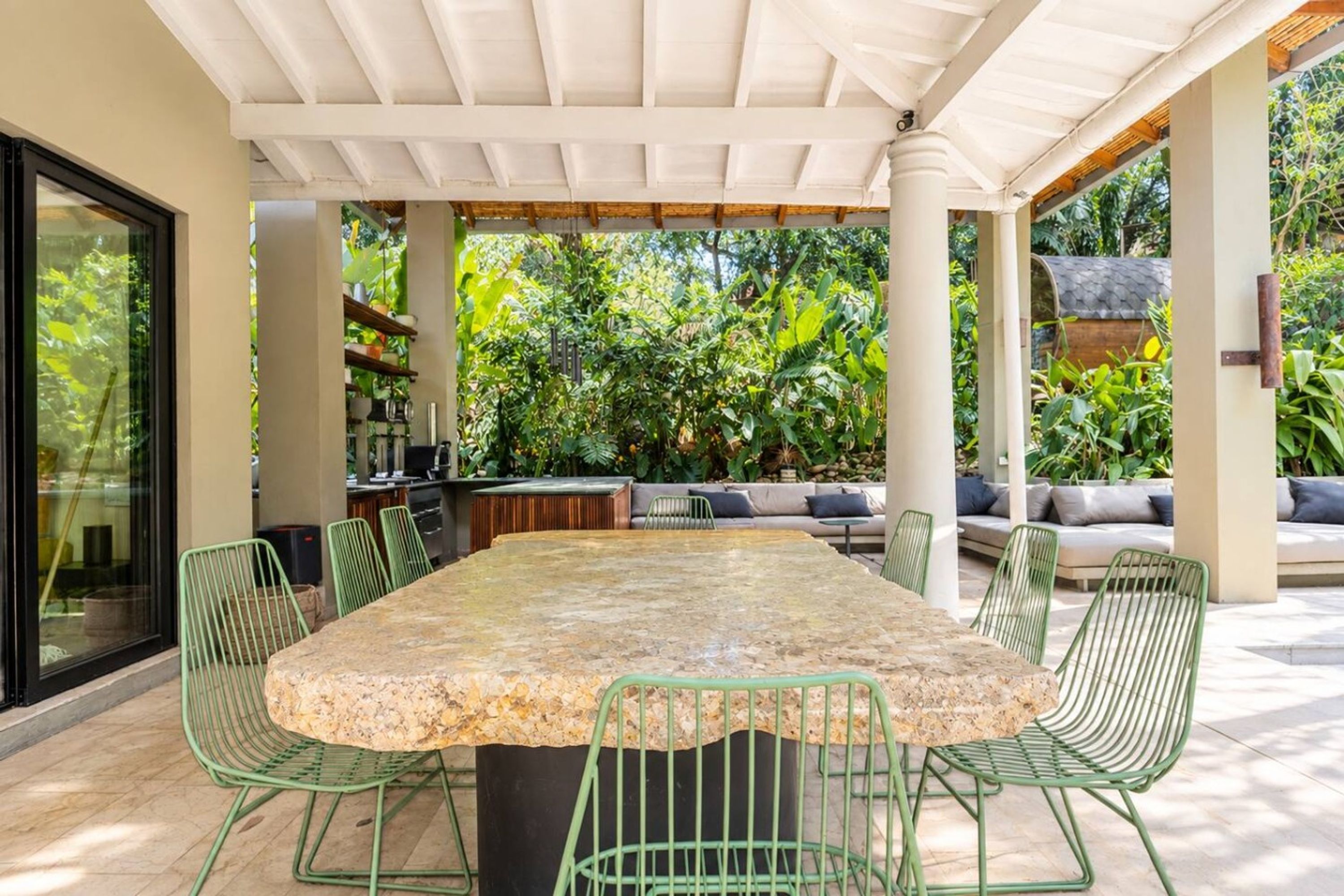 An outdoor dining area with a stone table and green chairs, surrounded by lush greenery and a covered seating area.