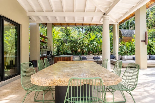 Un área de comedor al aire libre con una mesa de piedra y sillas verdes, rodeada de abundante vegetación y una zona de estar cubierta.