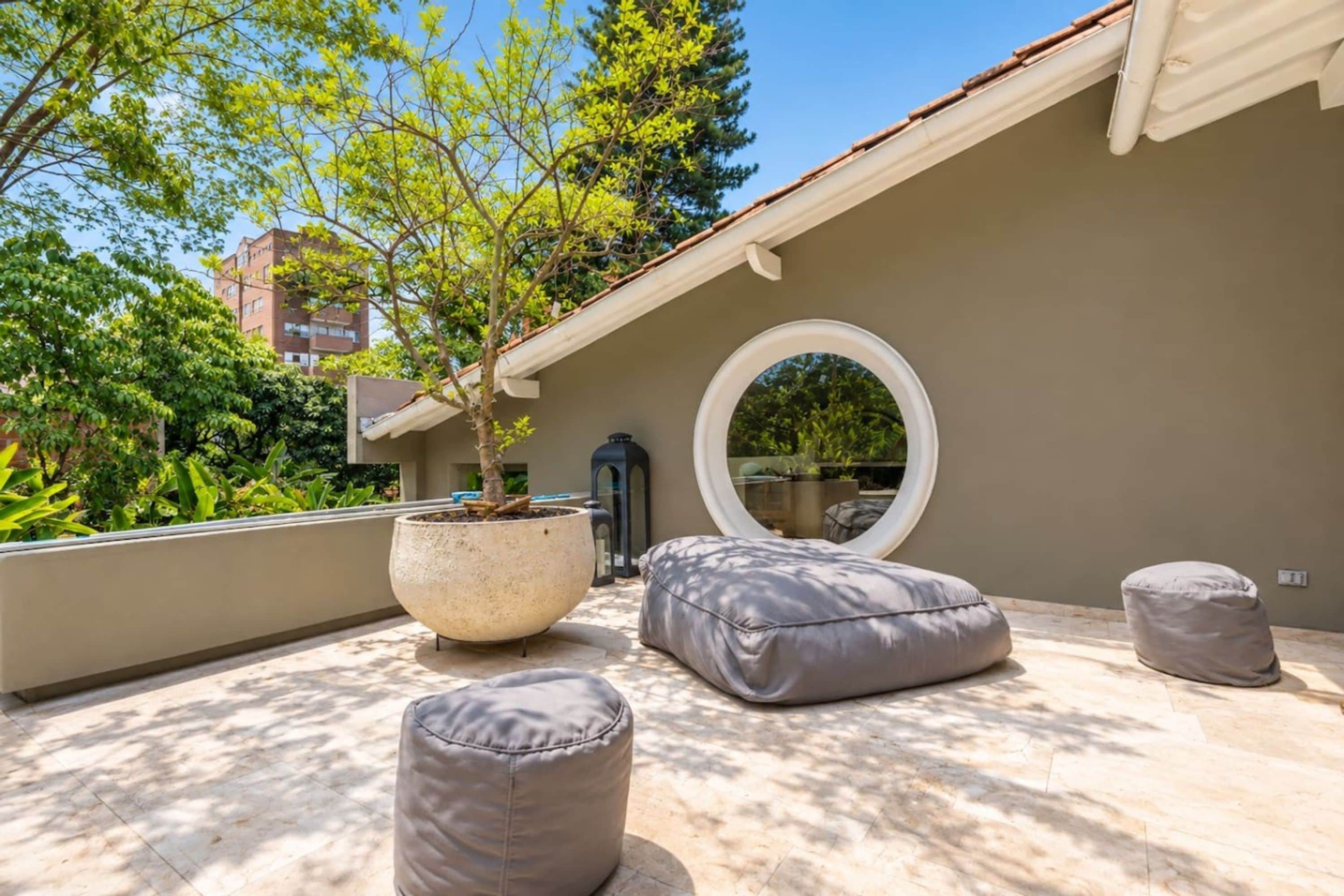 A rooftop patio with a potted tree, two grey poufs, and a round mirror.