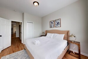 A hotel room with a bed, bedside tables, a nightstand, a lamp, a wall-mounted light fixture, and two framed pictures. - The Grandstand - Modern 4Br 3Ba Penthouse near Sox Stadium (Chicago)