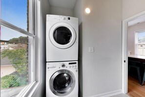 A laundry room with two stacked washing machines, a window with a view of a residential area, and a door leading to another room. - The Grandstand - Modern 4Br 3Ba Penthouse near Sox Stadium (Chicago)