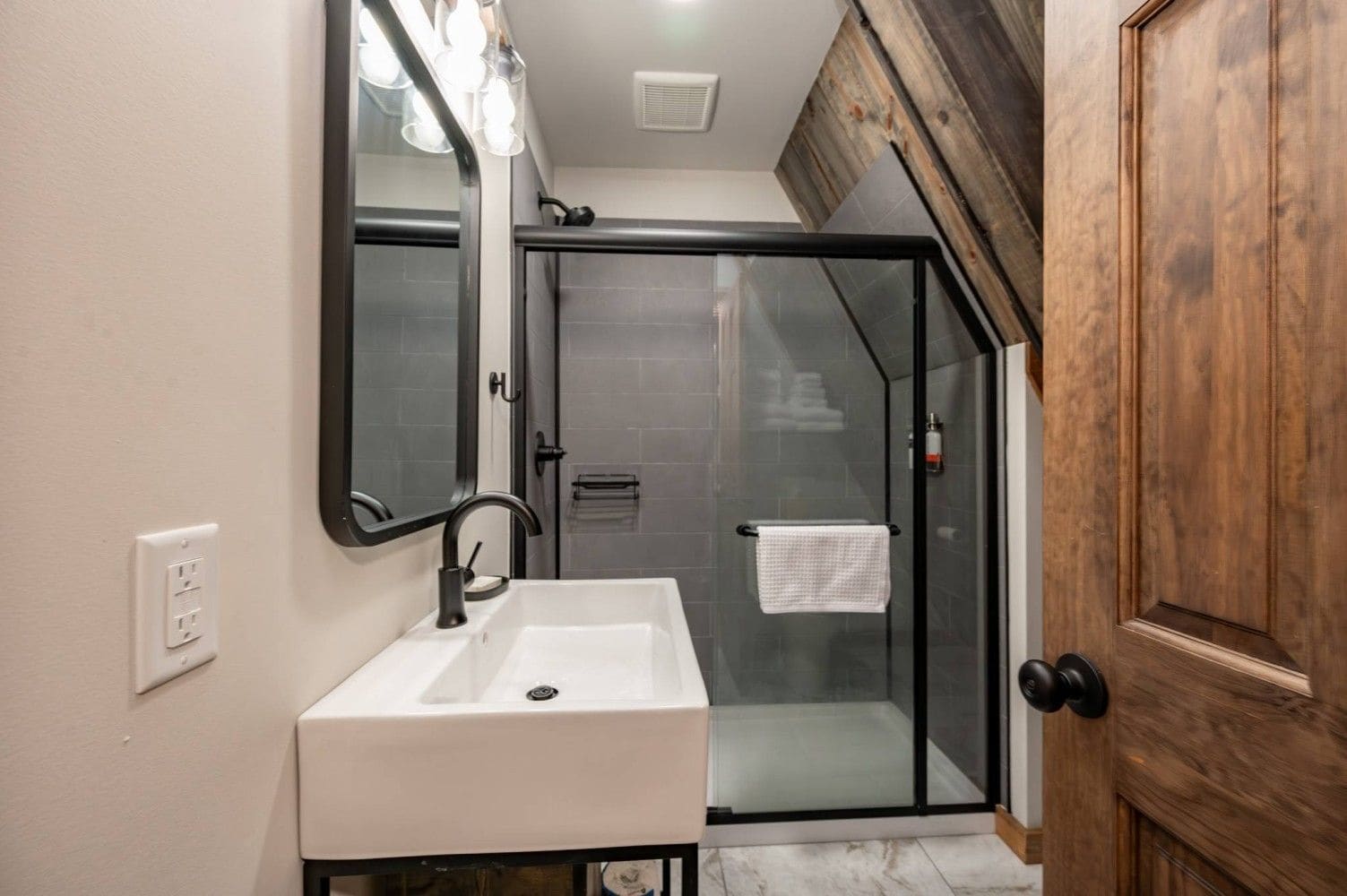 Un baño moderno con un lavabo blanco, cabina de ducha de vidrio y puerta de madera.