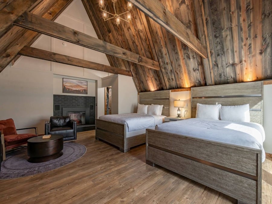 A loft-style bedroom with exposed wooden beams, two beds, a fireplace, and a seating area with a leather couch and a round table.