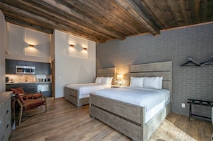 Quarto de hotel com teto de madeira, parede de tijolos, cama com roupa de cama branca, pequena cozinha e área de estar.