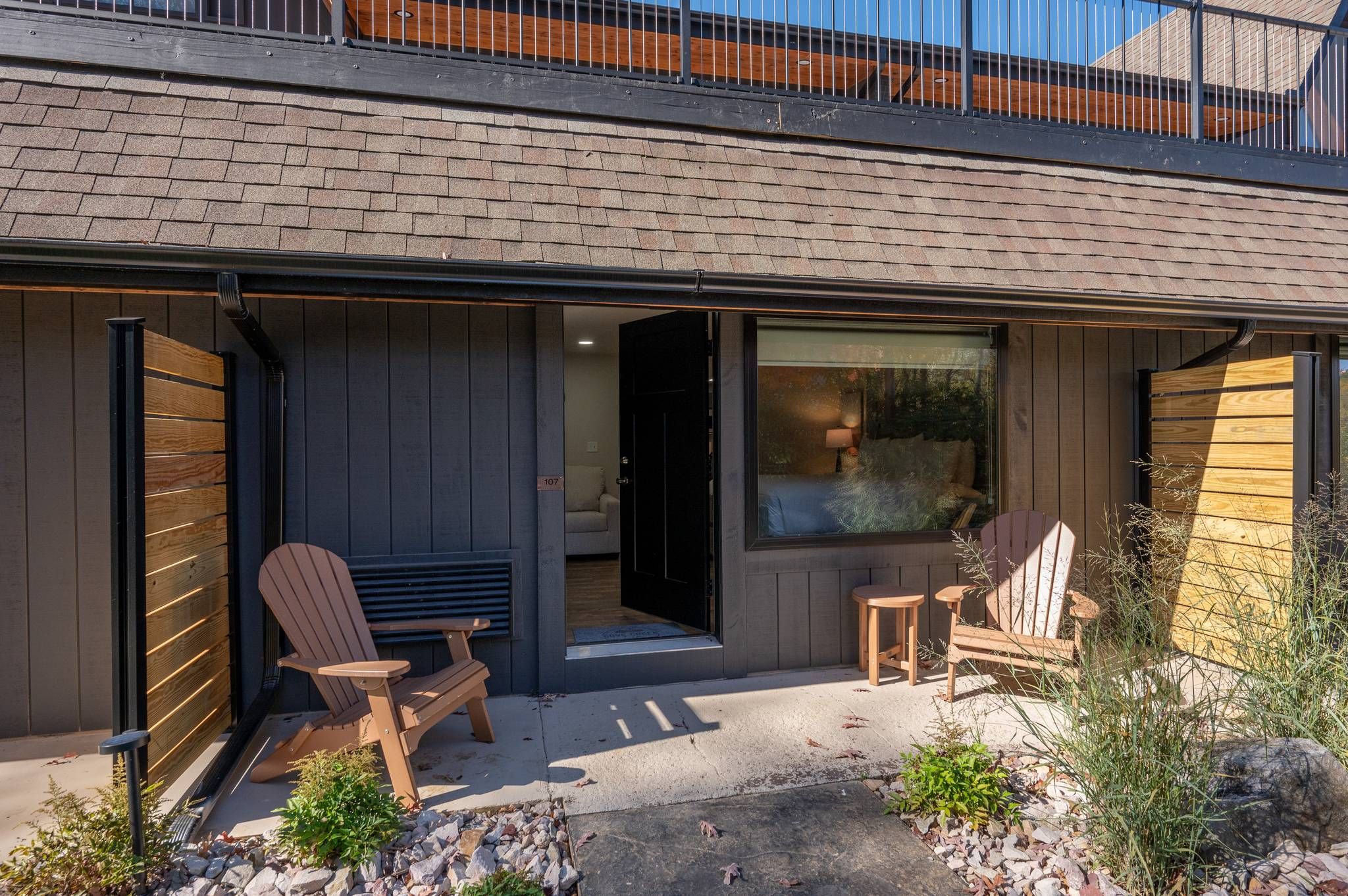 Ett modernt hus med en täckt uteplats, två Adirondack-stolar, ett litet bord och ett stort fönster.