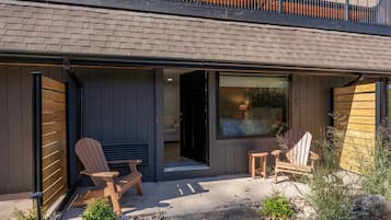 Ett modernt hus med en täckt uteplats, två Adirondack-stolar, ett litet bord och ett stort fönster.