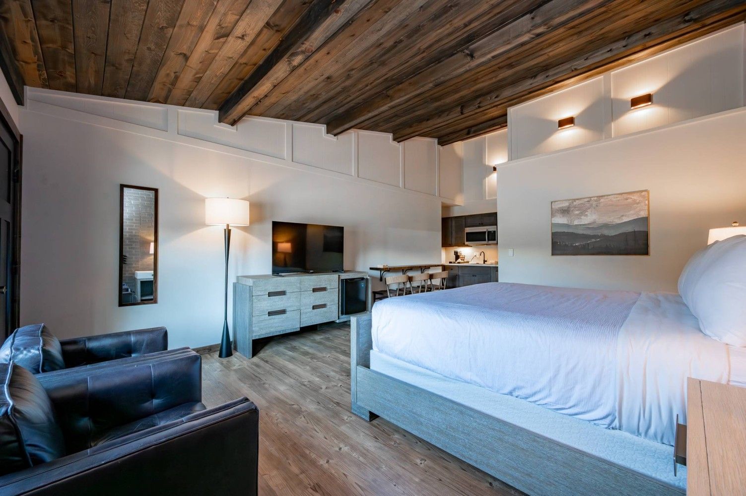 A modern hotel room with a large bed, a desk, a TV, and a kitchenette.