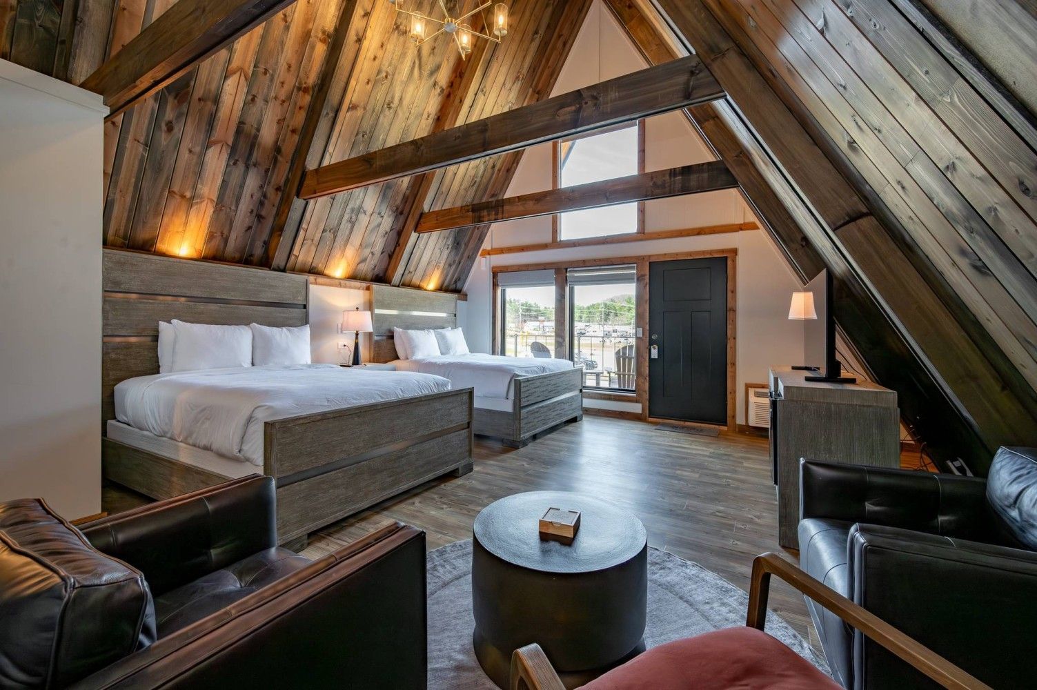 A spacious loft bedroom with a high ceiling, wooden beams, a large bed, a smaller bed, a sofa, a round coffee table, and a TV.