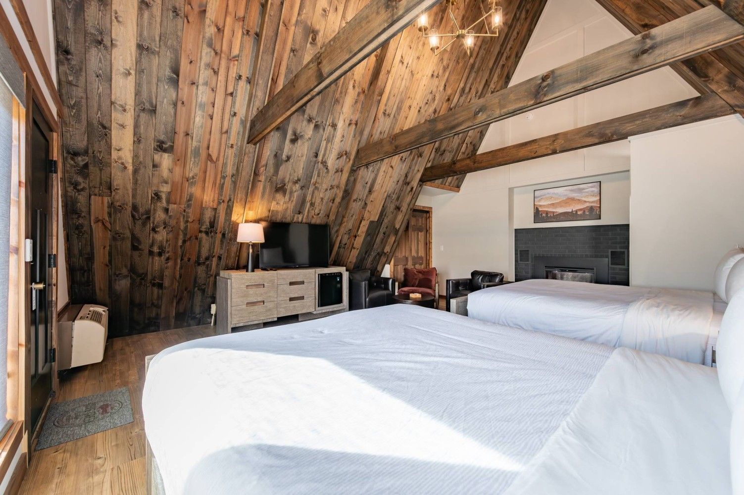 Quarto com teto inclinado, vigas de madeira, uma cama, uma televisão, lareira e um quadro na parede.