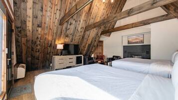 A bedroom with a sloped ceiling, wooden beams, a bed, a TV, a fireplace, and a painting on the wall.