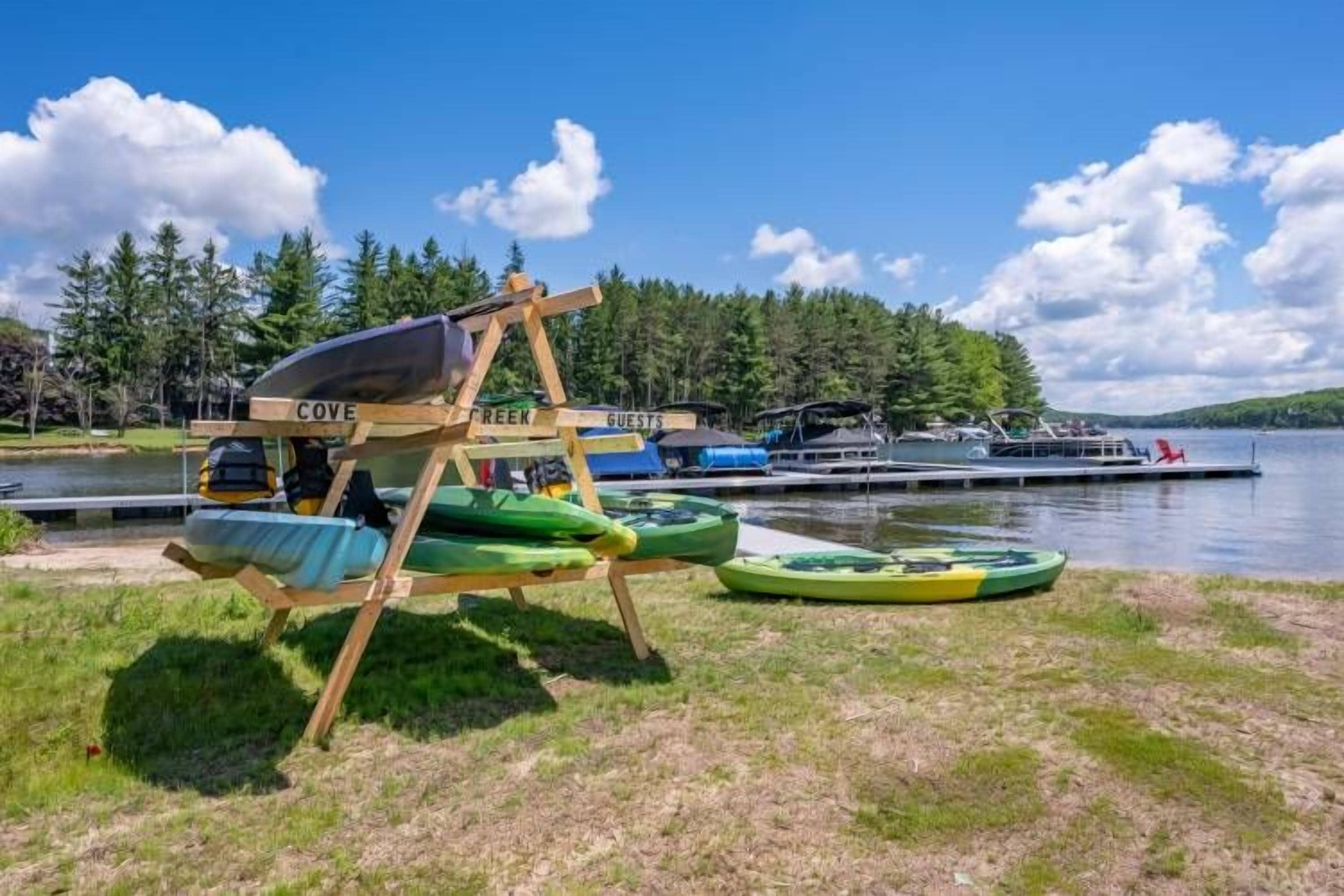 Een landschap aan het meer met een houten constructie waarop 'COVE' en 'GUESTS' staan, bedoeld voor kamperen, omgeven door boten en een bebost gebied.