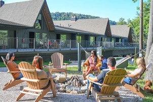 Um grupo de pessoas sentadas em cadeiras Adirondack ao redor de uma fogueira em ambiente externo, com um chalé ao fundo.