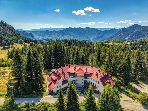 A multi-story building with a red-tiled roof surrounded by dense forest, set against a backdrop of mountains.