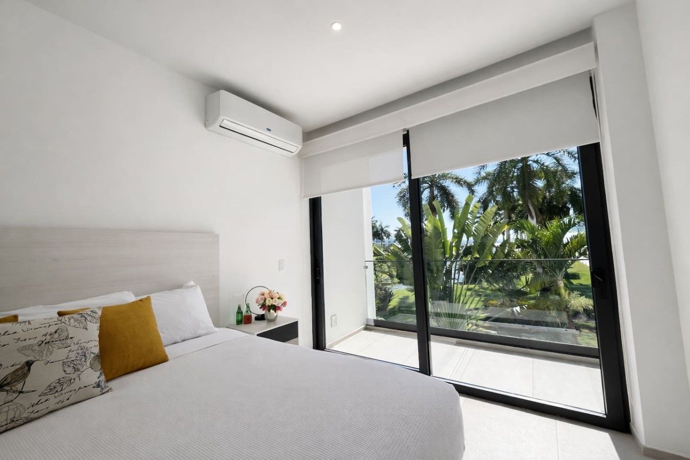 Un dormitorio moderno con una cama grande, una mesita de noche, una pequeña mesa con un jarrón y un balcón con vistas a palmeras.