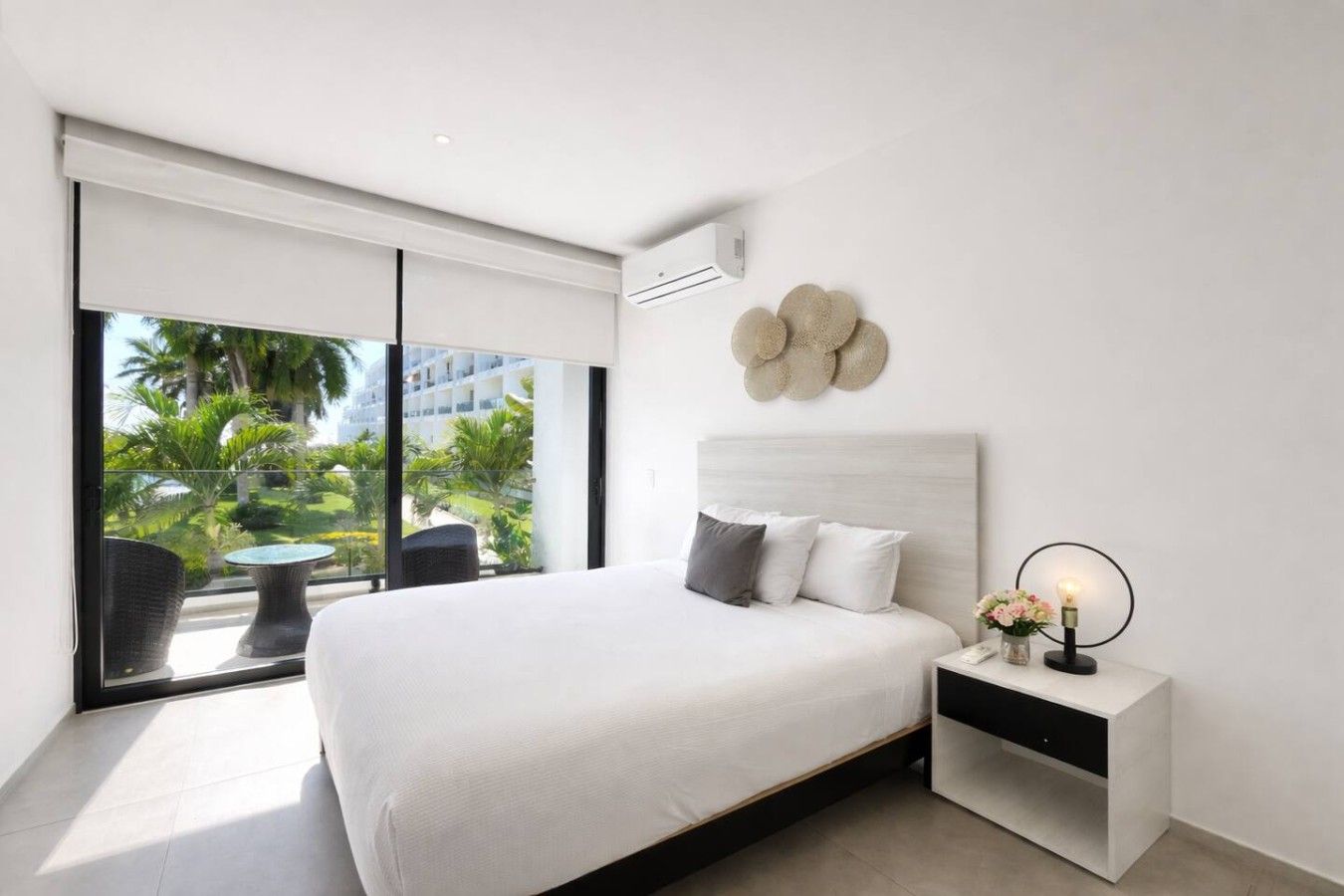 A modern bedroom with a large bed, a nightstand, a wall-mounted air conditioner, and a view of palm trees through a sliding glass door.
