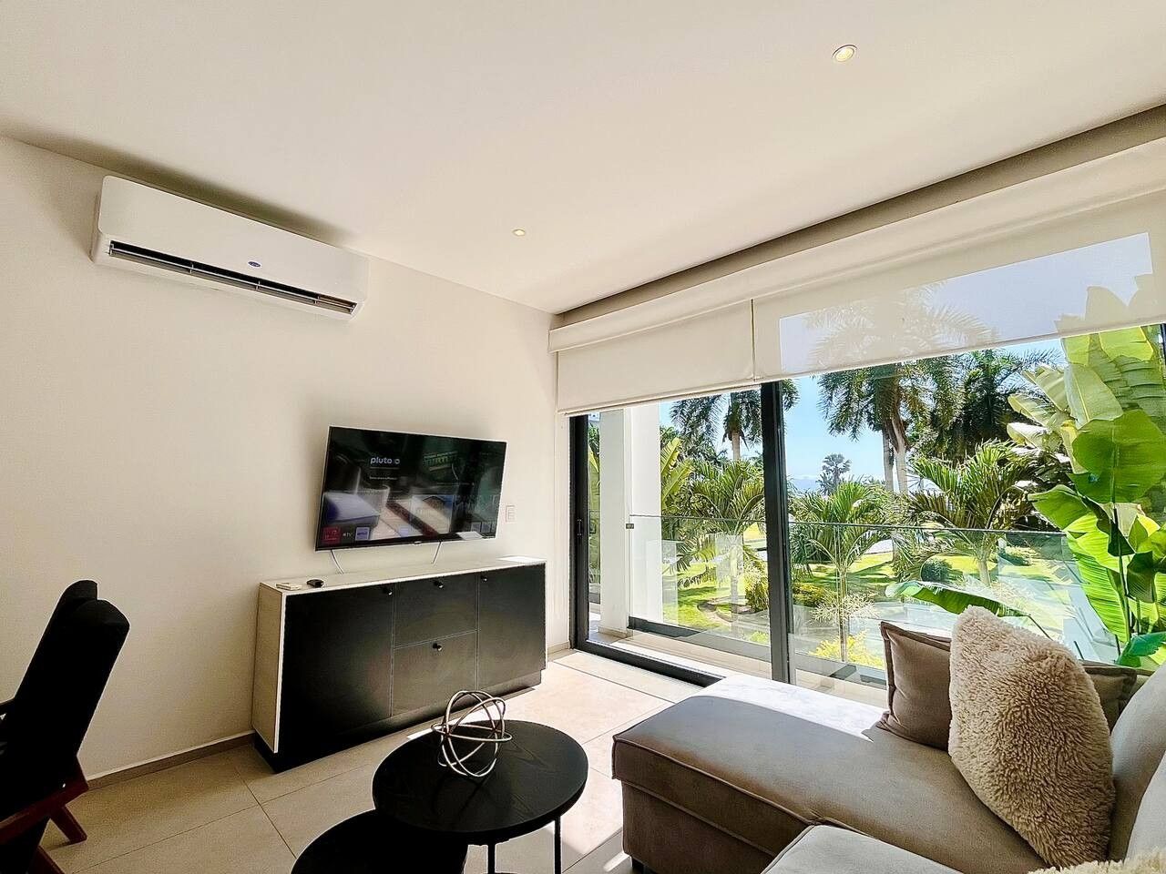 A modern living room with a flat-screen TV, a sofa, a coffee table, and a sliding glass door leading to a balcony with a view of greenery.