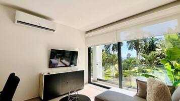 A modern living room with a flat-screen TV, a sofa, a coffee table, and a sliding glass door leading to a balcony with a view of greenery.