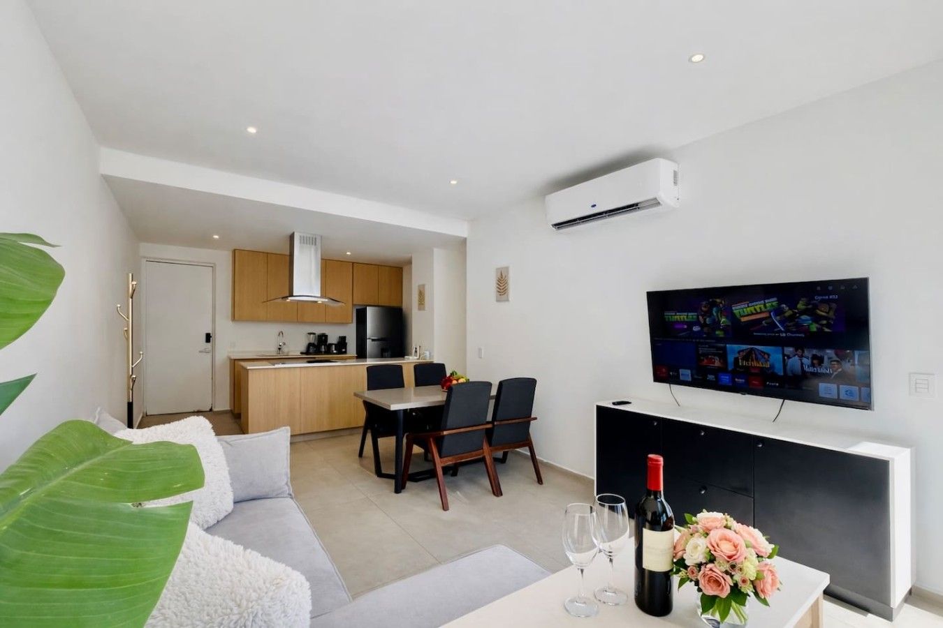 A modern living room with a sofa, a dining area with chairs, a kitchen with a fridge, and a TV mounted on the wall.