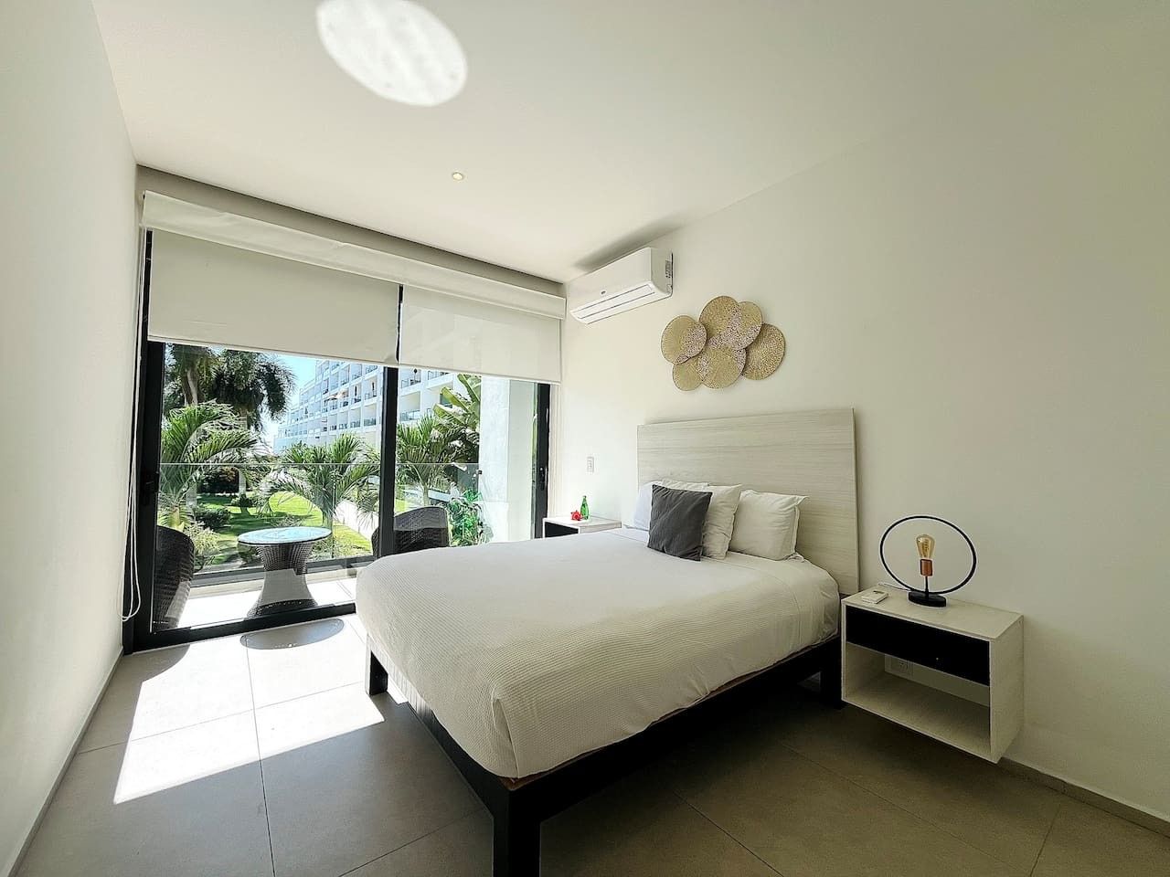 Une chambre moderne avec un grand lit, une table de chevet avec une lampe, et une vue sur un balcon agrémenté d’une table et de chaises.