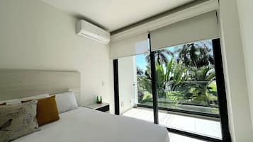 A bedroom with a large bed, a nightstand, a window with a view of greenery, and an air conditioning unit.