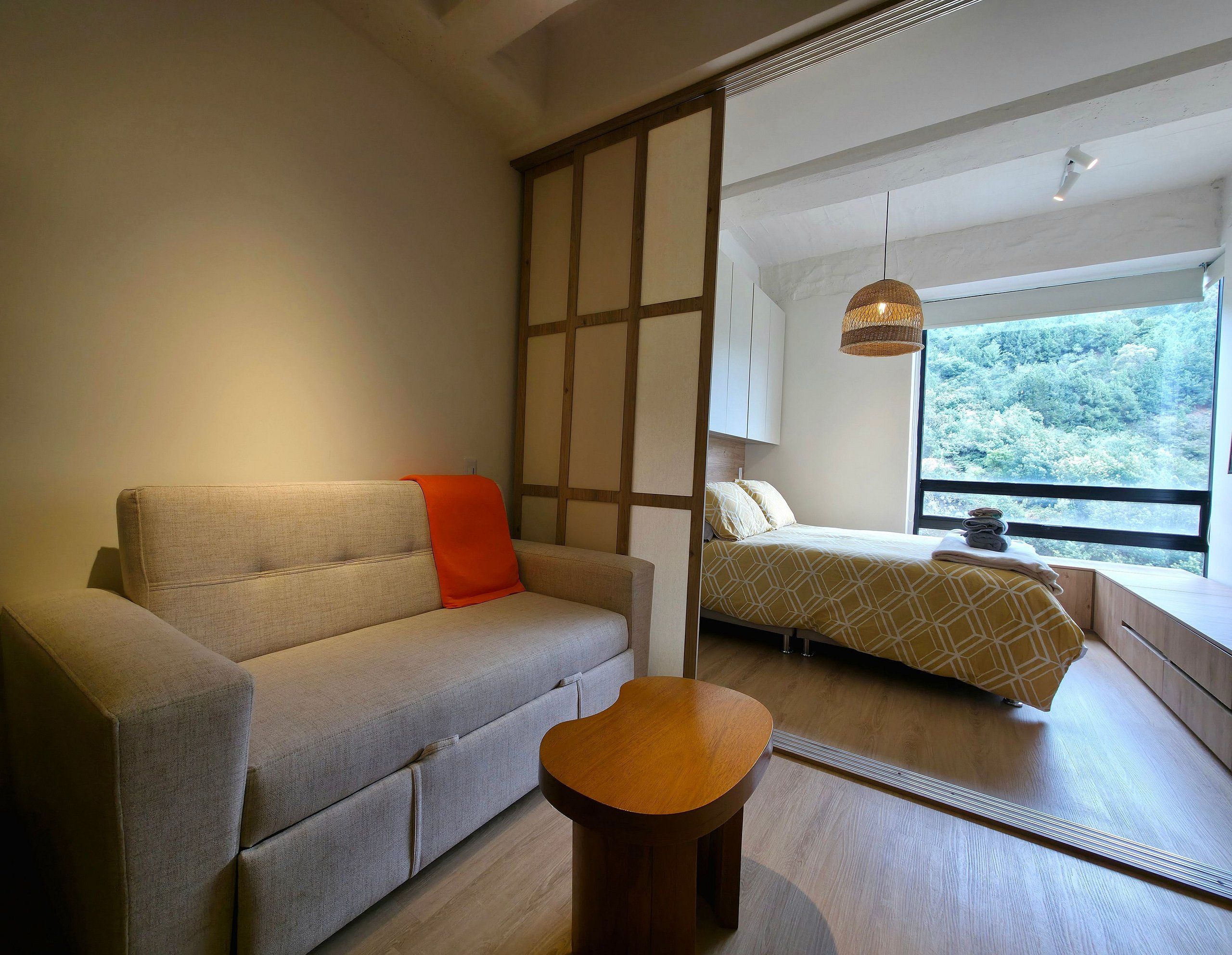 A modern room with a sofa, a small wooden stool, a bed with a geometric patterned bedspread, and a view of a forest through a window.