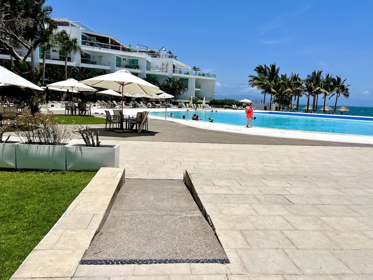 Área da piscina do resort com vista clara para o oceano, cadeiras de descanso e guarda-sóis.