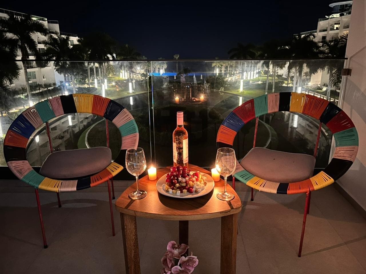 Mesa de jantar com duas cadeiras, uma garrafa e um prato de comida, com vista para uma paisagem urbana através de um guarda-corpo de vidro.