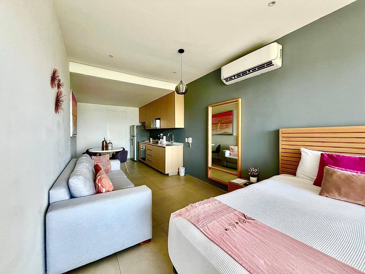 A modern hotel room with a sofa, a bed with a pink blanket, a small kitchen area, and a large mirror.