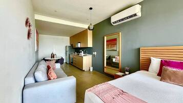 A modern hotel room with a sofa, a bed with a pink blanket, a small kitchen area, and a large mirror.