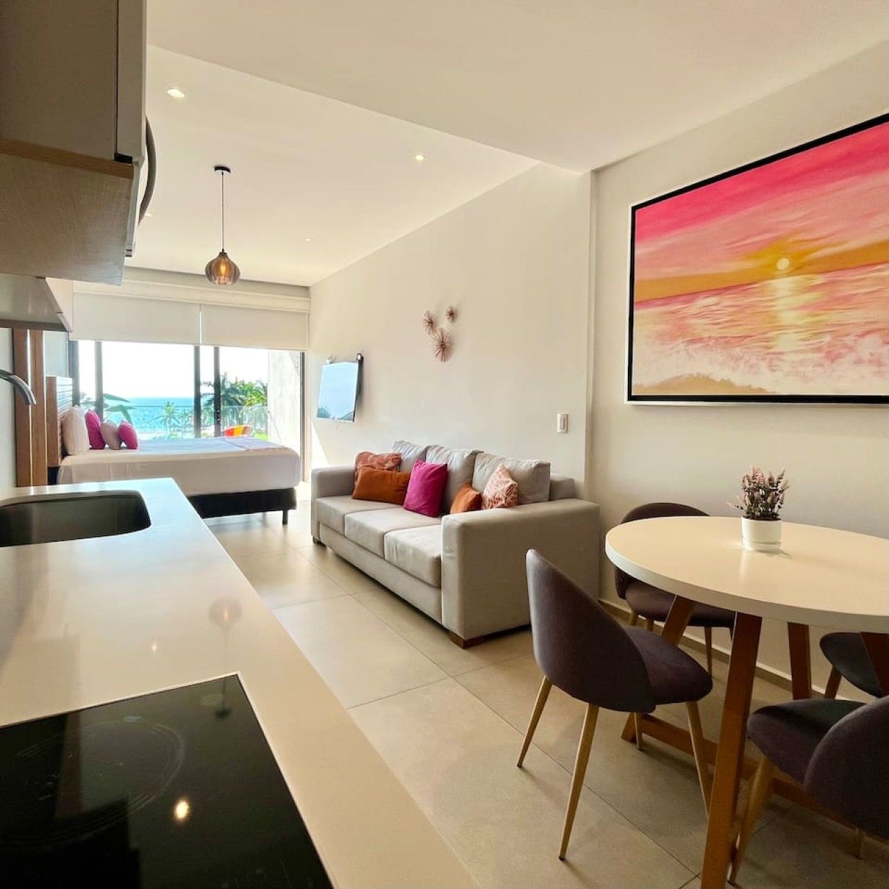 A modern living space with a kitchen area, a dining table with chairs, a sofa with cushions, and a large painting on the wall.
