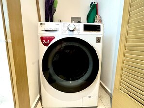 A front-loading LG Direct Drive 12kg washer with a 2-enveloper capacity, located in a laundry room with a wooden door and a light-colored wall. - Comodo Penthouse Aria1012 Playa/Albercas Nvo. Vta. (Nuevo Vallarta)
