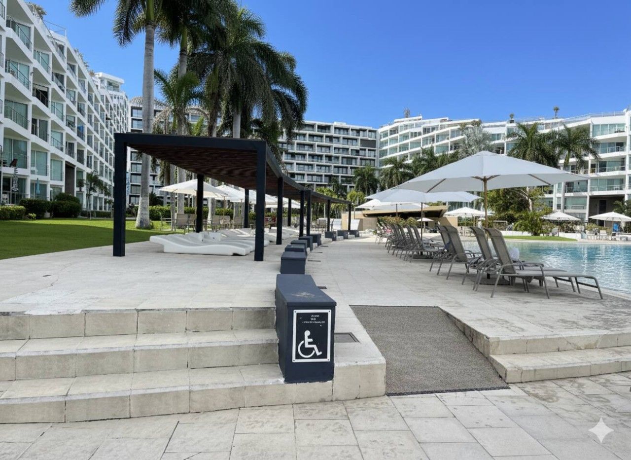 Un espace aménagé au bord de la piscine, comprenant un coin salon couvert, des chaises longues et un panneau indiquant l’accès pour les personnes handicapées.
