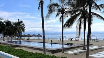 Un área de piscina con palmeras, una playa y un cielo despejado.