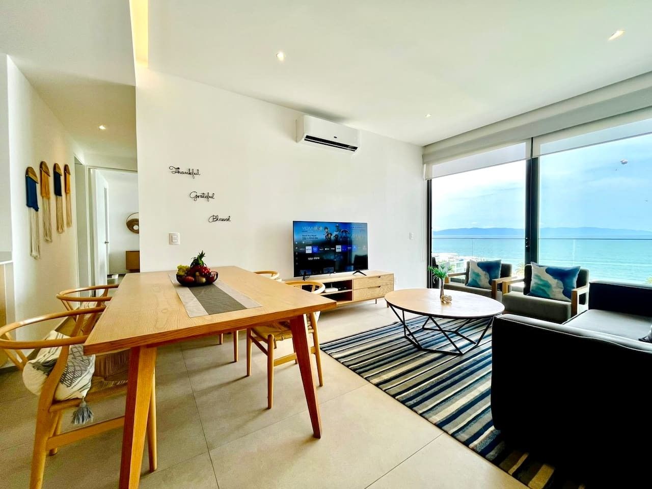 A modern living room with a dining area, a flat-screen TV, and a view of the sea.