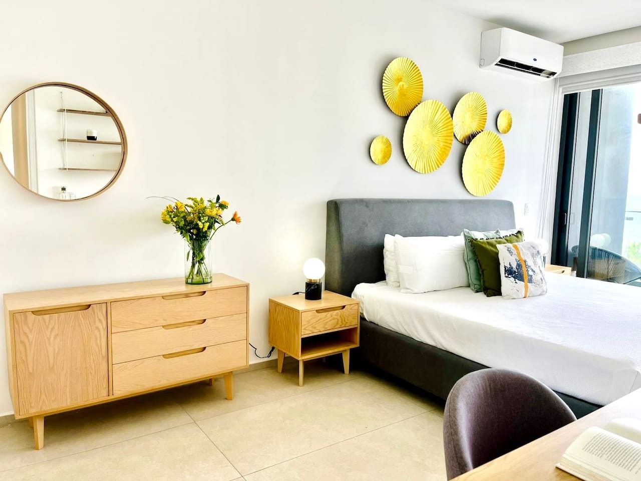 A modern hotel room with a bed, bedside table, nightstand, dresser, and a large window.